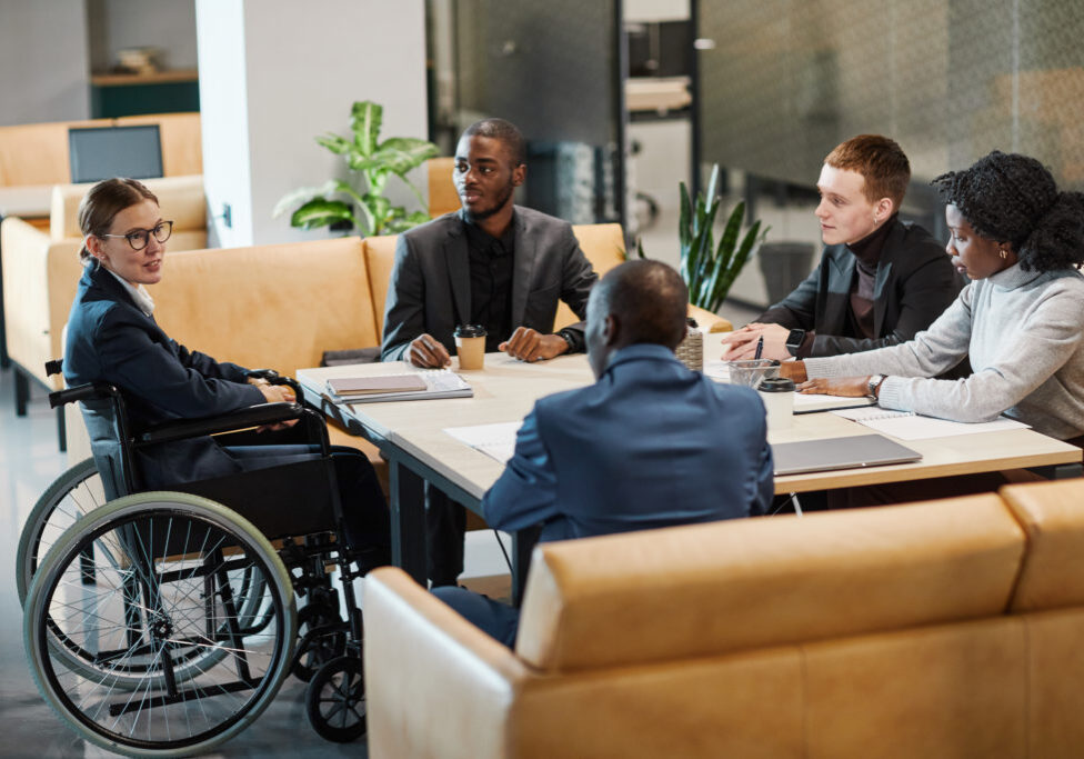 Full length portrait of successful businesswoman using wheelchair at meeting and talking to colleagues in modern office space